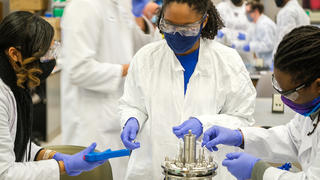 3 black women in lab PPE taking careful measurements while working with a small pressure vessel