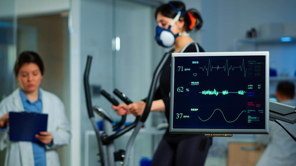 Monitor showing EKG scan of athlete running on cross trainer and medical specialists supervise exercise in background, controlling physical activity, measuring heart rate in sports science laboratory