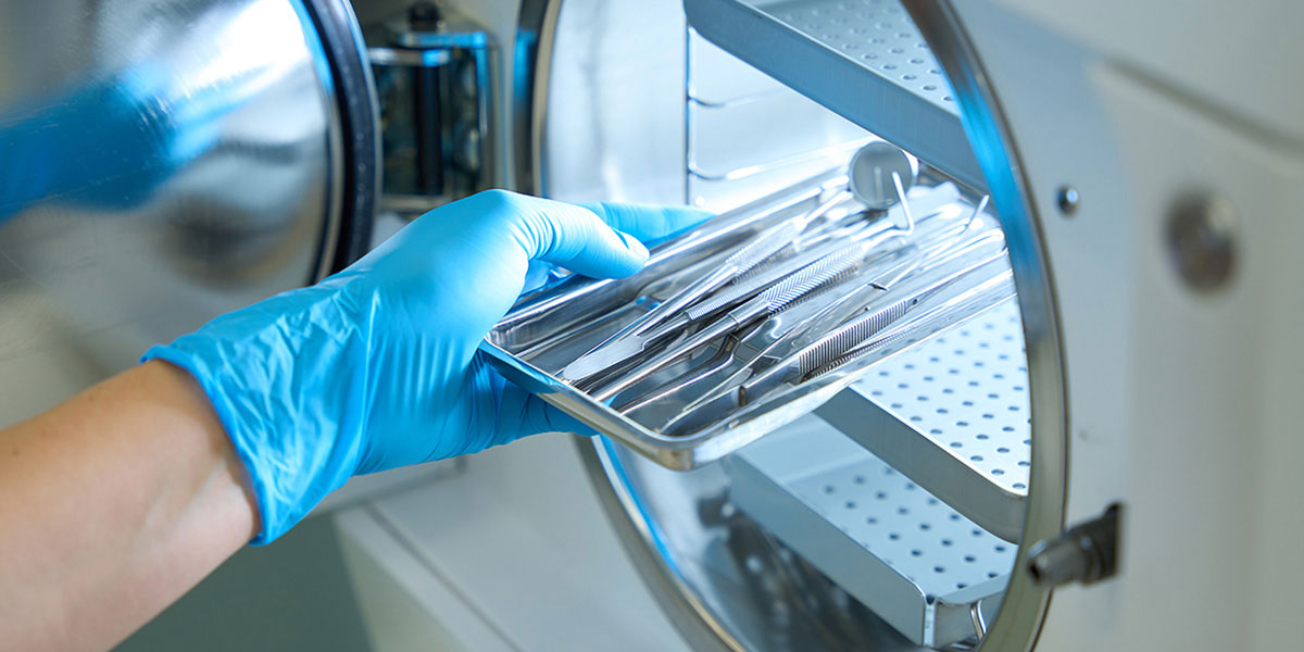 A gloved hand places a tray of sterilized dental tools into an open autoclave.
