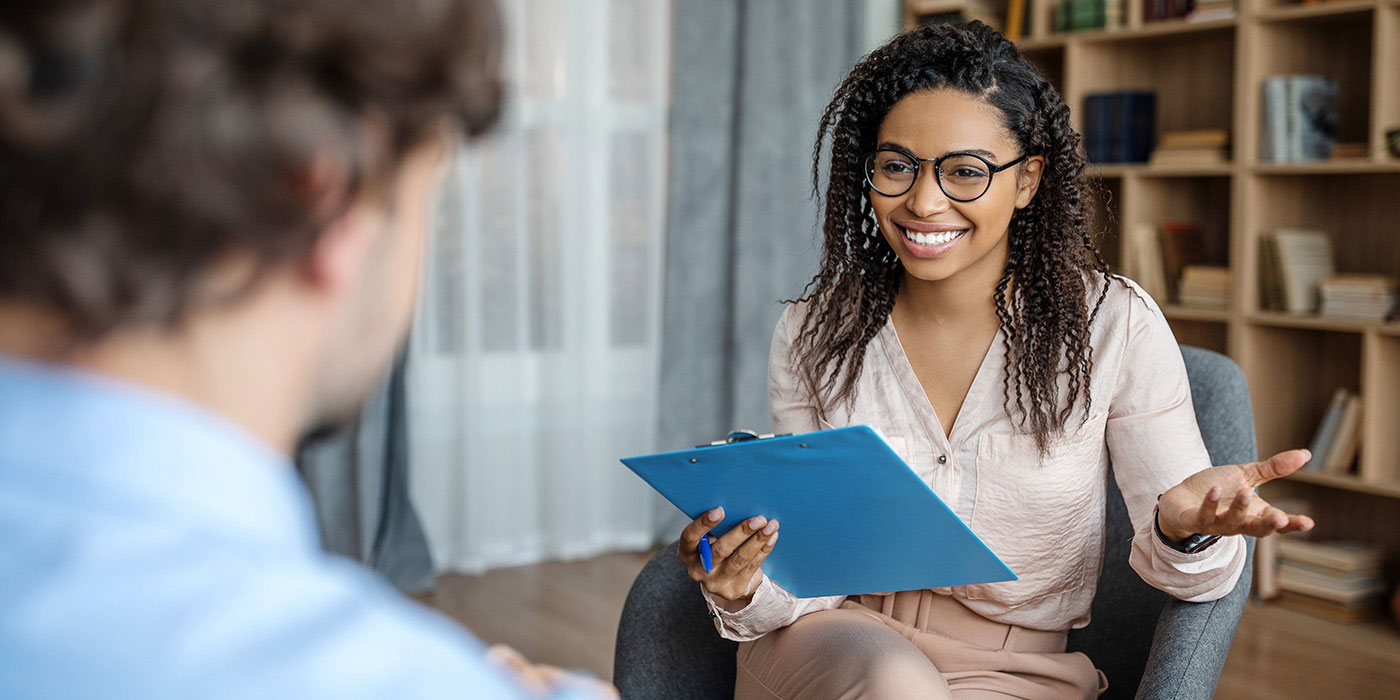  A smiling female therapist wearing glasses and a light-colored blouse holds a clipboard and gestures while speaking to a male client whose back is to the camera.