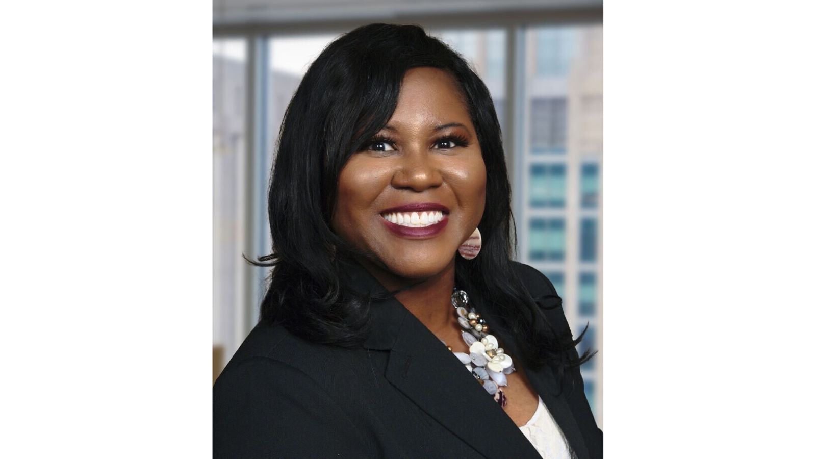 Professional headshot of Dena J. King smiling in a black blazer and statement necklace against a blurred office background.