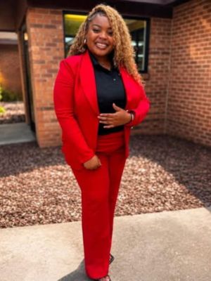 Kianna Jones smiling outdoors in a red pantsuit and black top in front of a brick building.