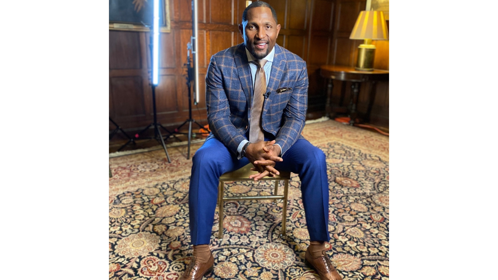 Professional portrait of Ray Lewis seated in a wood-paneled room, wearing a blue suit and tie, smiling with studio lighting equipment visible in the background.