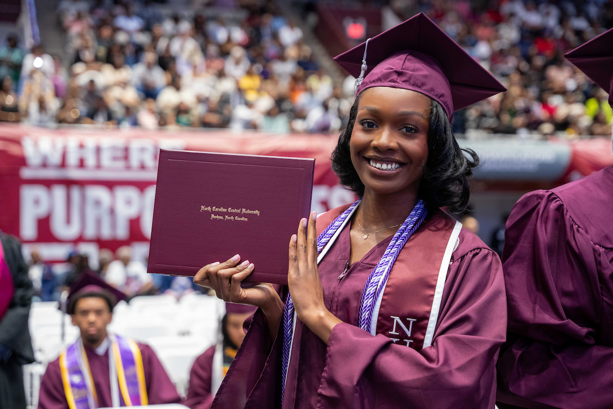 North Carolina Central University graduate in maroon cap and gown holds degree during commencement ceremony.