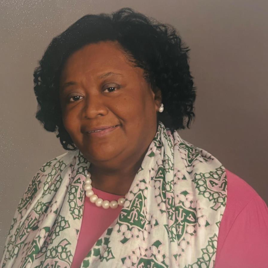 Portrait of a smiling woman wearing pearl jewelry and a patterned scarf against a neutral background.