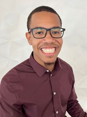 Professional portrait of Will smiling, wearing glasses and a maroon button-down shirt.