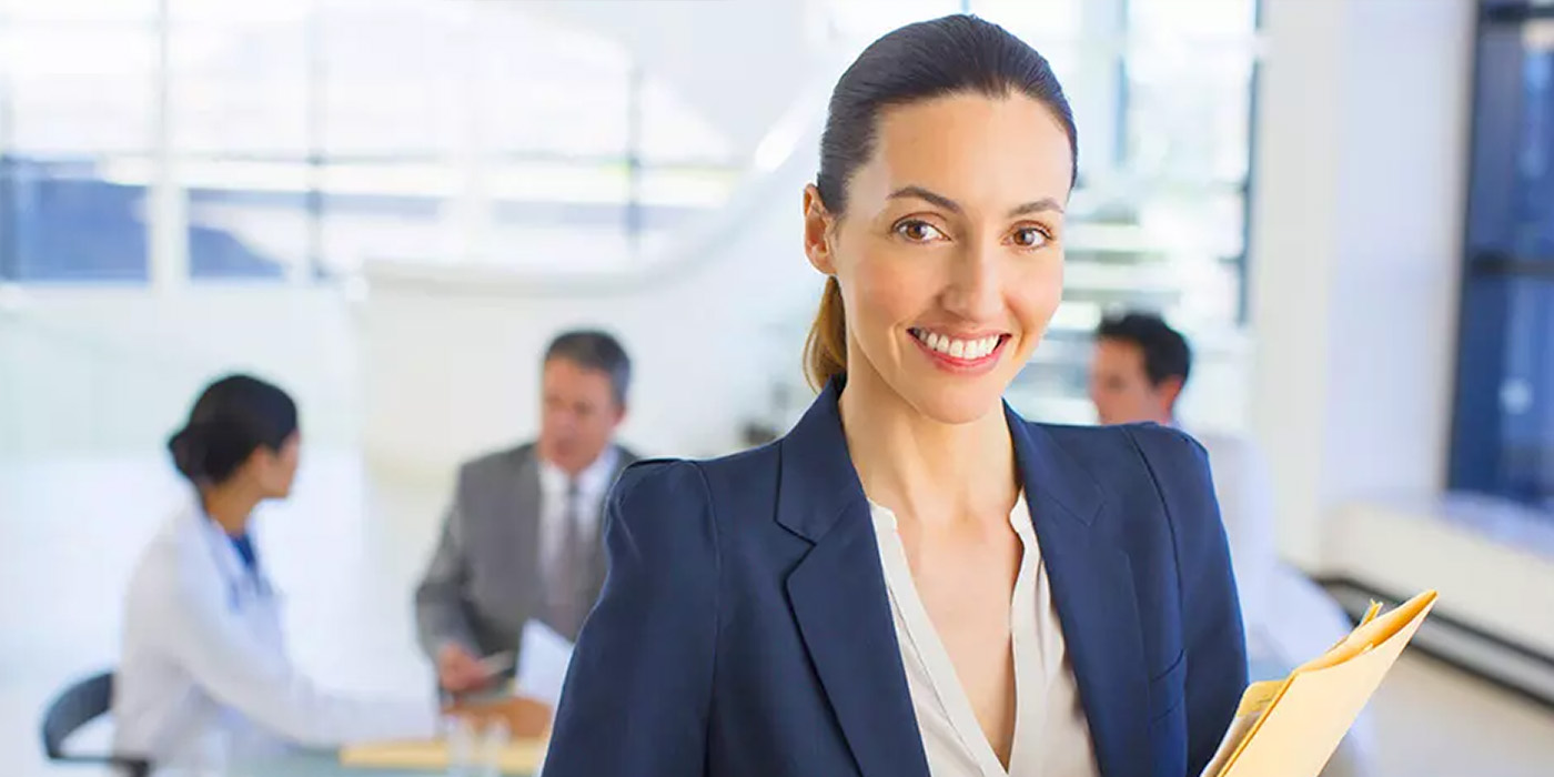  A smiling woman in a navy blazer and white blouse holds a manila folder, looking at the camera with three blurred figures in a meeting room in the background.