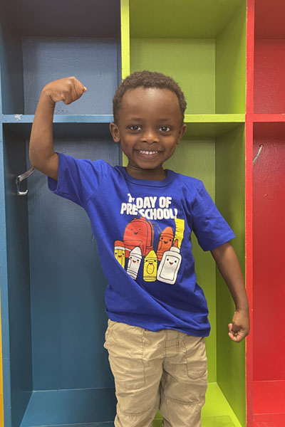 Child flexing his arm, wearing a "First Day of Preschool" t-shirt
