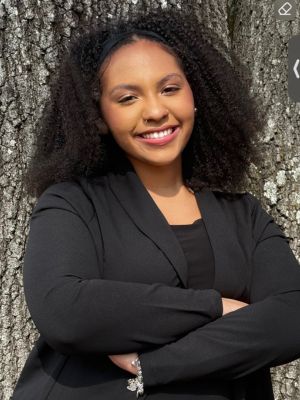 Professional portrait of Gabby smiling in a black blazer outdoors.