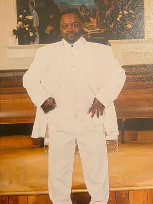 Portrait of a man wearing an all-white suit, standing confidently inside a church sanctuary with wooden pews and religious artwork in the background.