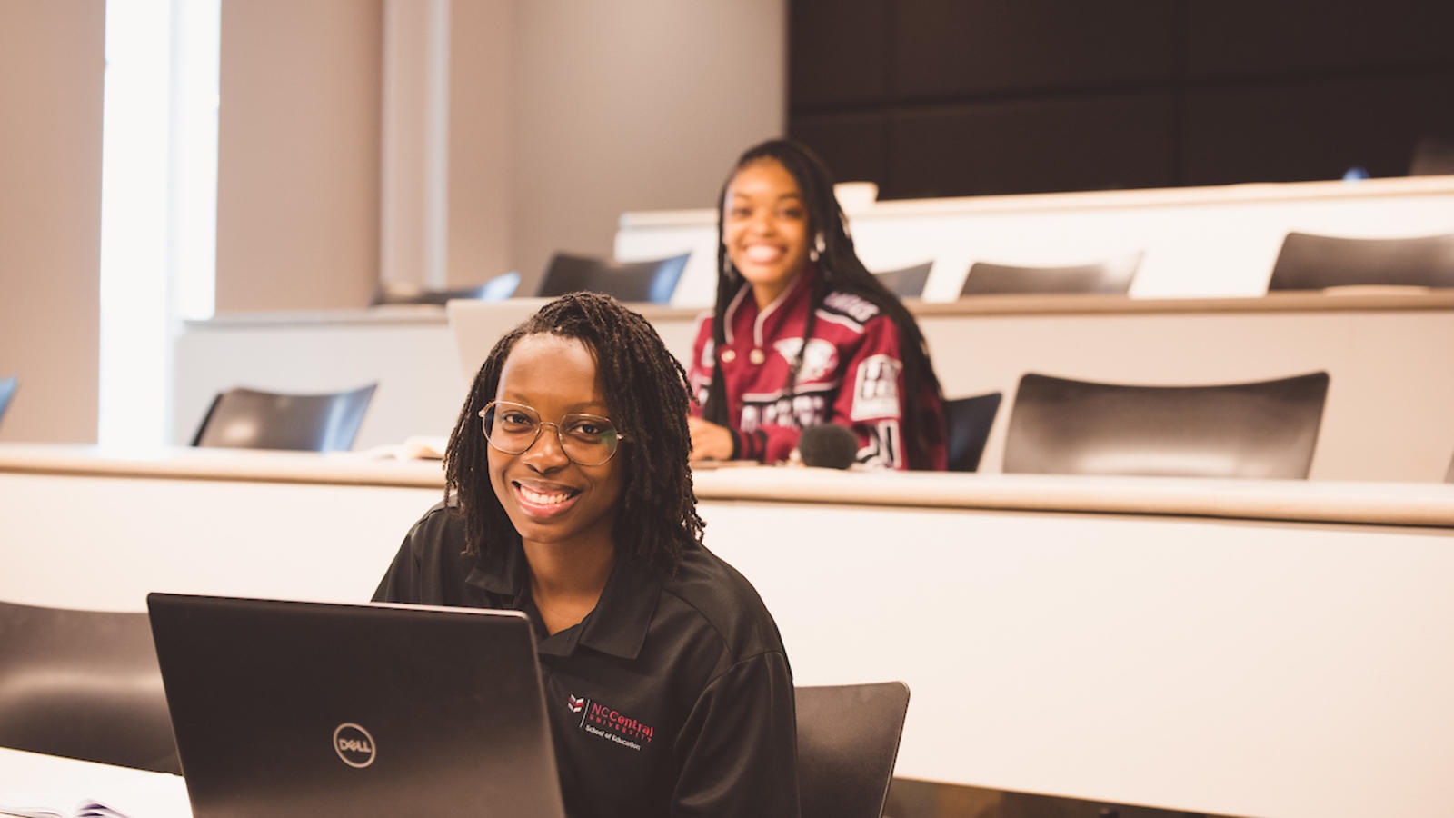 Two smiling students sitting in a lecture hall and looking towards the camera