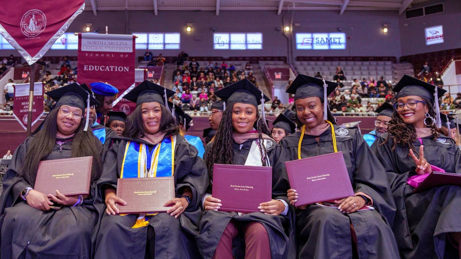 Graduates in caps and gowns holding diplomas, smiling at a ceremony.