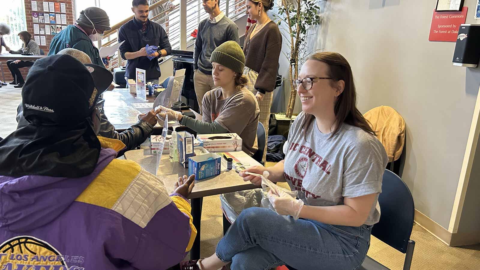 A smiling woman with glasses and a gray T-shirt, wearing white gloves, sits at a table and helps a person in a purple and yellow jacket, who is also wearing gloves, at a health screening event.