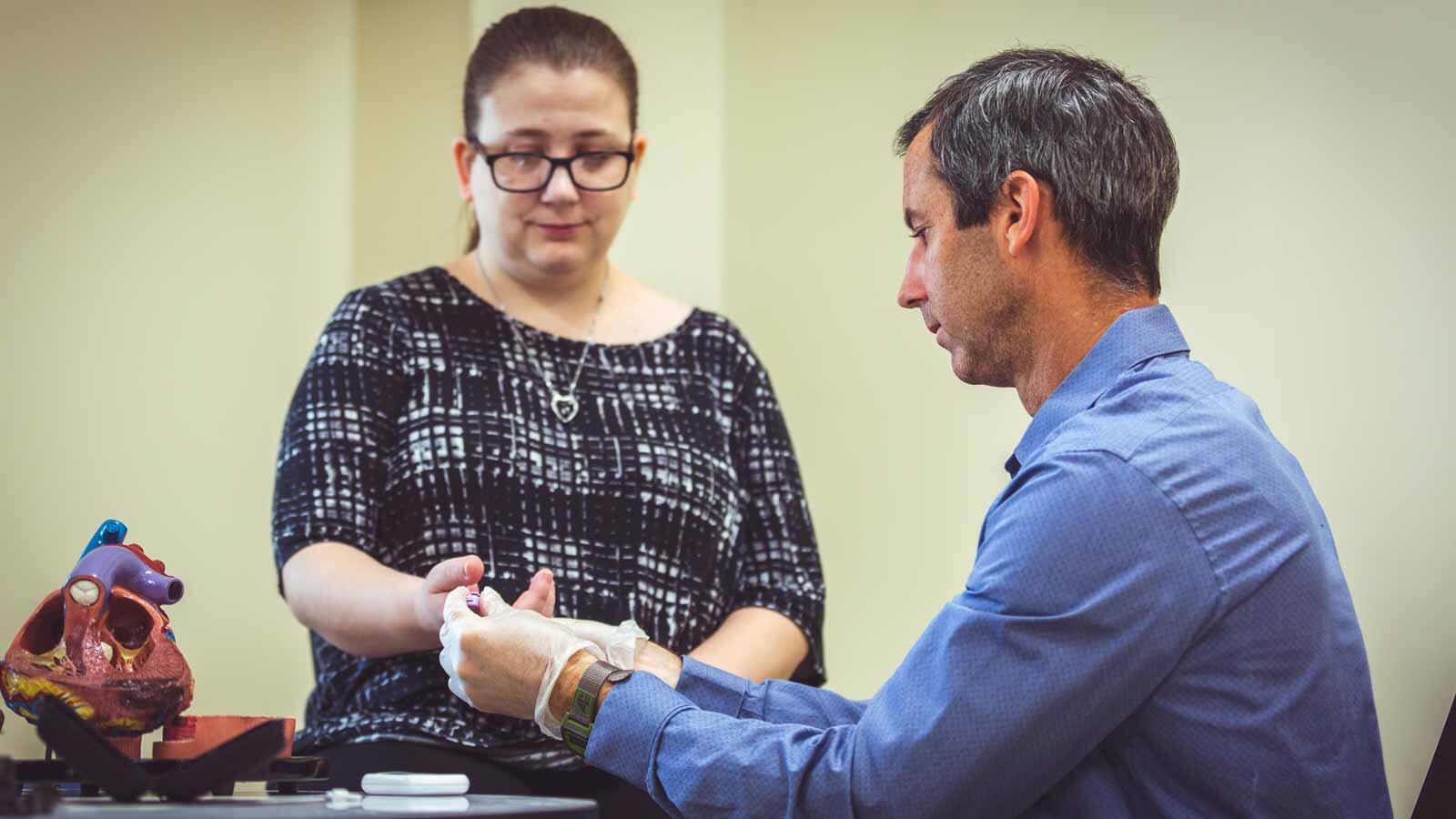 A medical professional wearing a blue shirt and white gloves is holding a patient's hand and pricking their finger with a lancet while a model of a human heart sits on the table to their left.