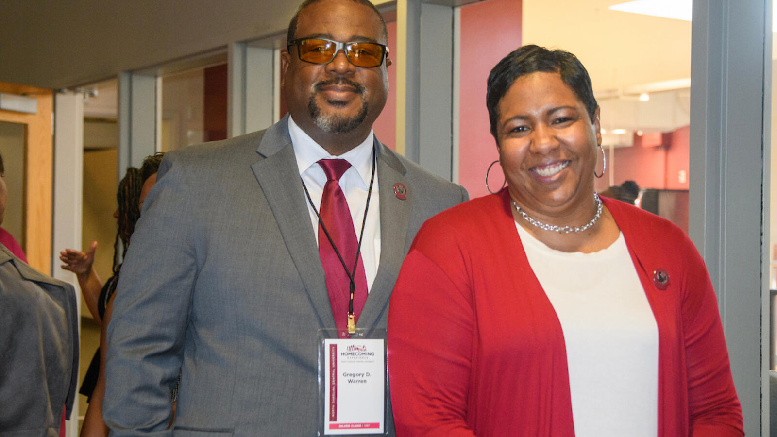 A man and woman smiling and standing together indoors. The man wears a gray suit with a red tie and name badge, and the woman wears a red cardigan over a white top.