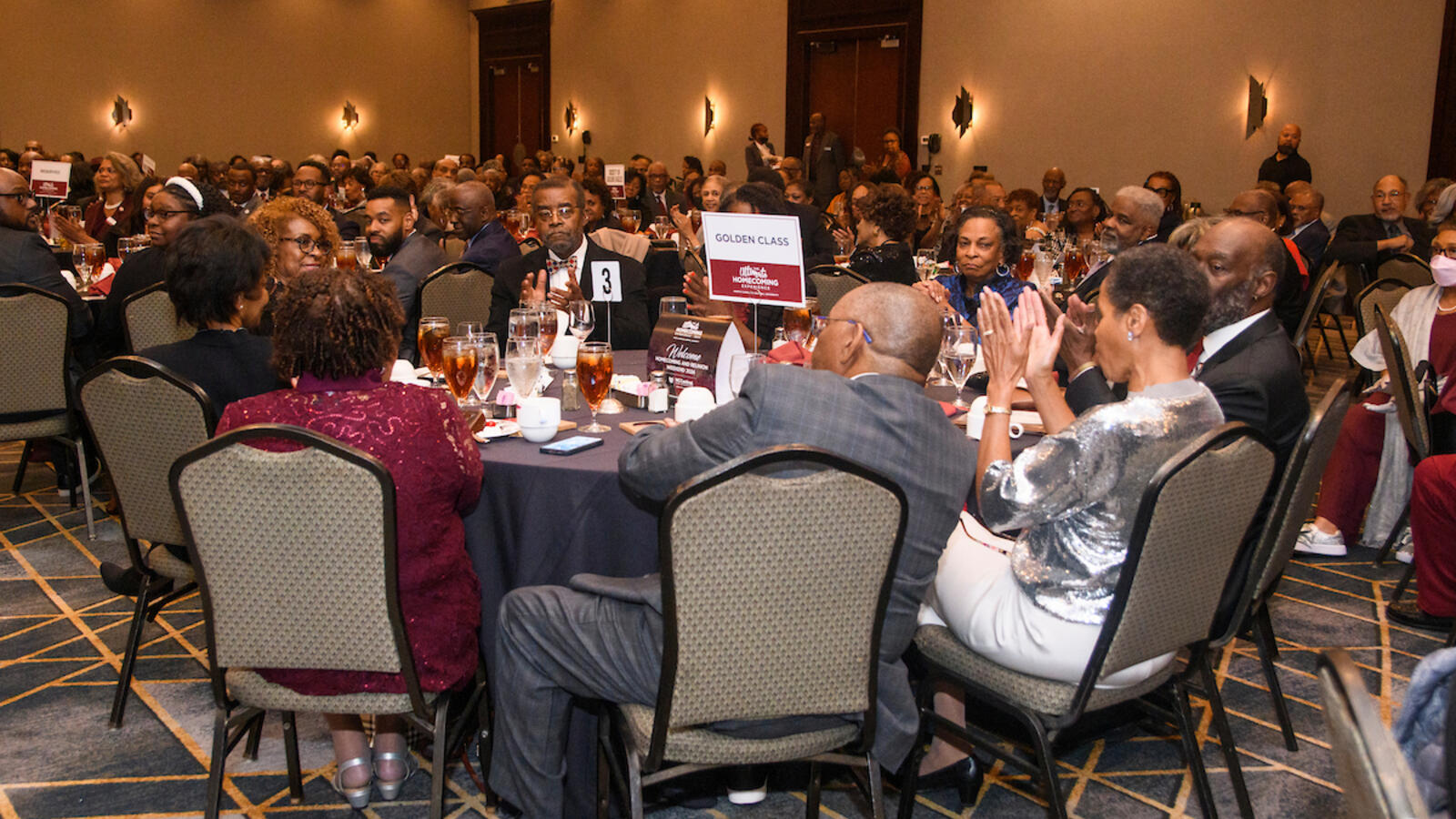 A large group of people dressed in formal attire sit at round tables during a banquet event, with a sign reading “Golden Class” displayed at one table.