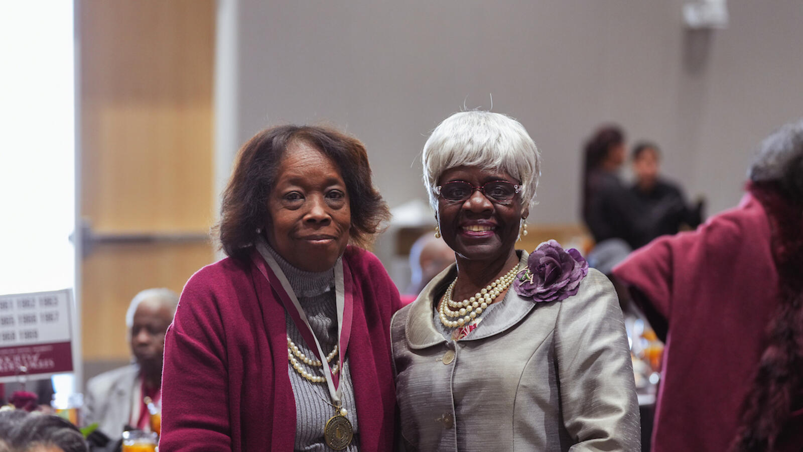 Two older women smile while posing together at a formal luncheon. One wears a maroon sweater with a medal, and the other wears a silver suit with pearls and a purple flower accessory.