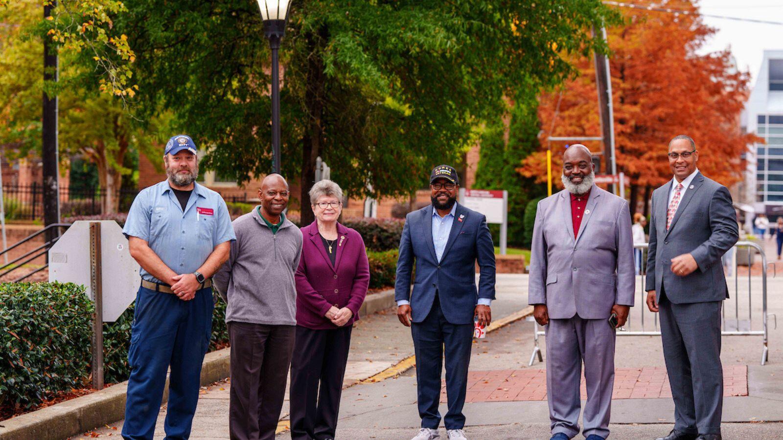 Group of six people, including veterans and university staff, stand outdoors on a campus walkway surrounded by fall foliage during a Veterans Day event at North Carolina Central University.