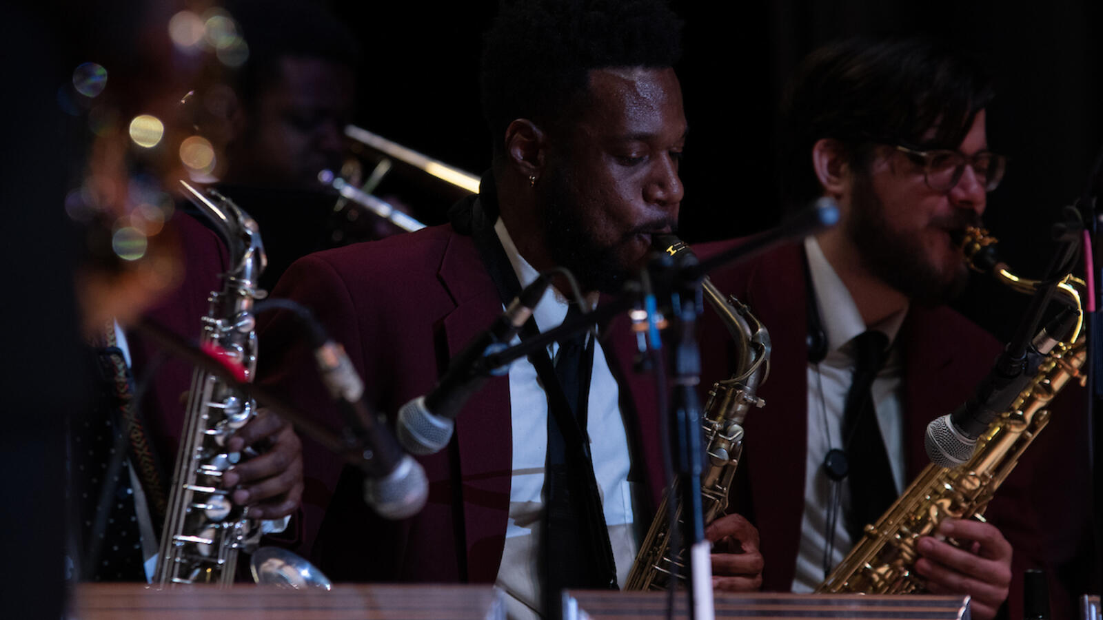 Jazz musicians perform on stage wearing maroon blazers and black ties, playing saxophones and brass instruments during a live concert.