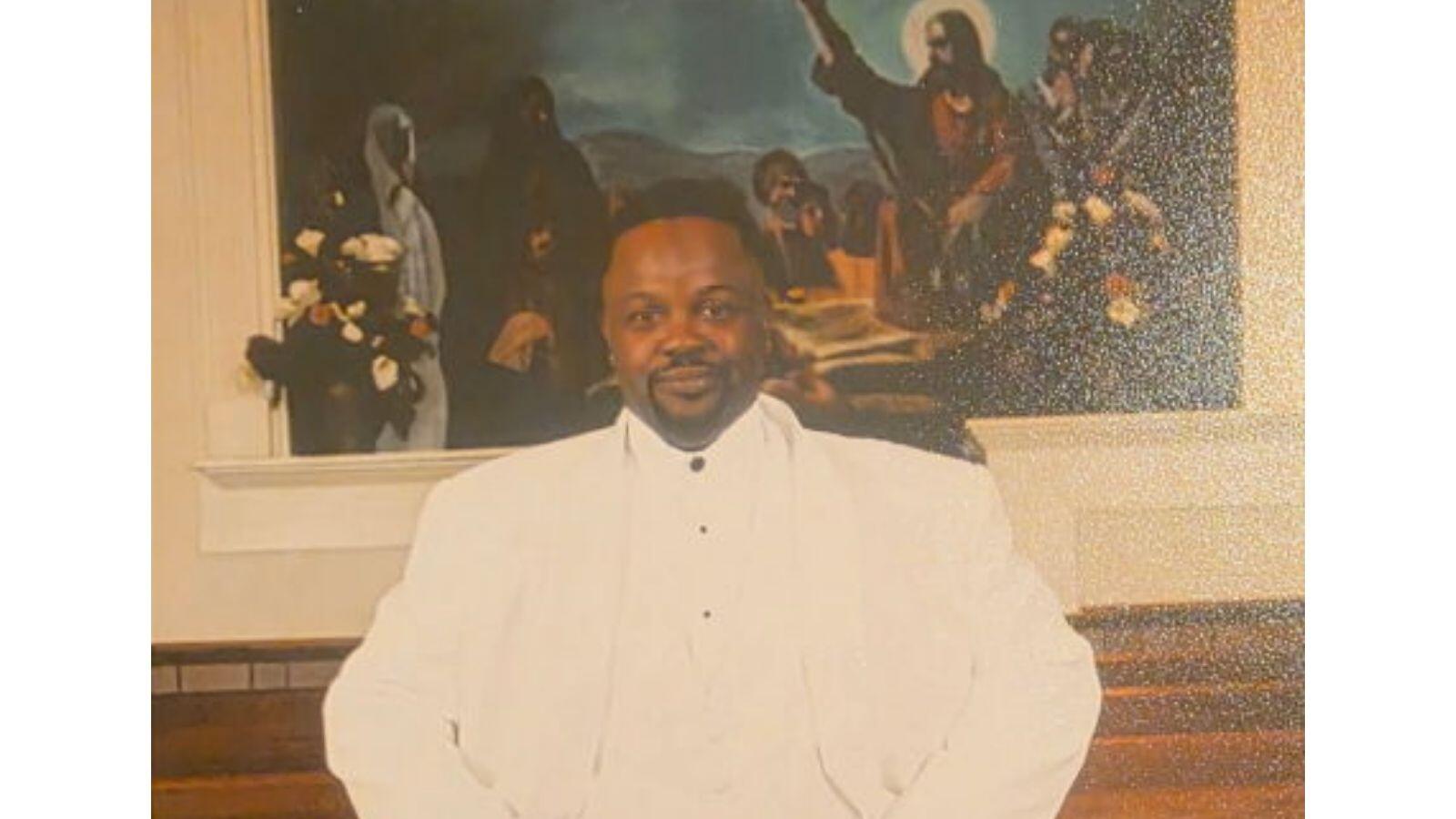 Portrait of Mr. James Winston wearing an all-white suit, standing confidently inside a church sanctuary with wooden pews and religious artwork in the background.