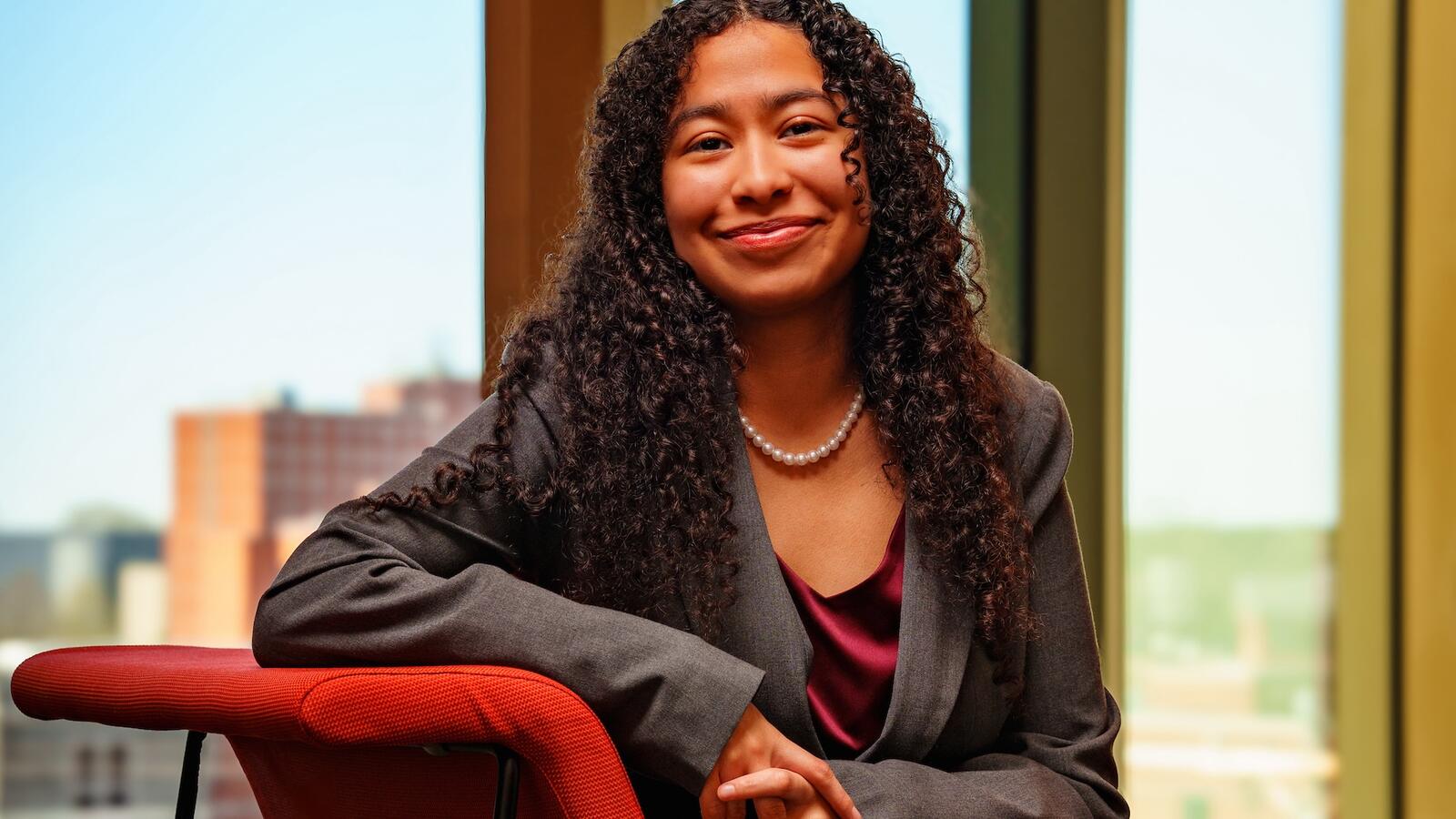 Angie Melchor-Roque sitting on red chair in gray suit