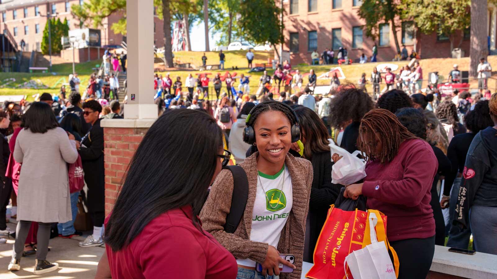 A smiling woman wearing over-ear headphones and a John Deere shirt stands outdoors amidst a large crowd at a college campus event.
