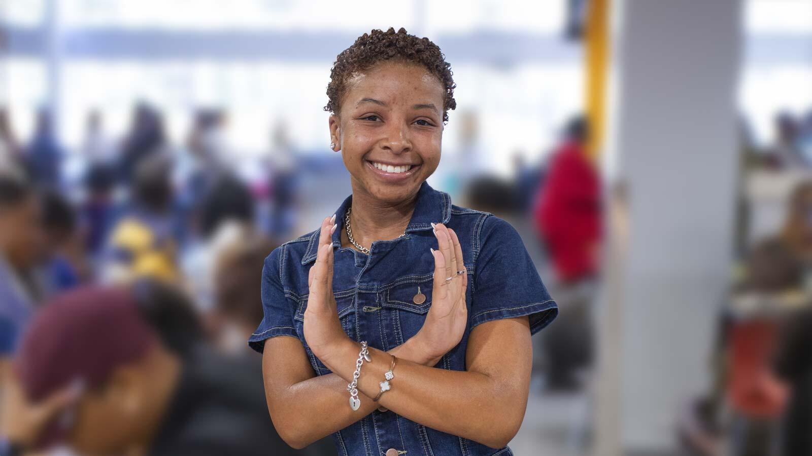 A smiling young woman in a denim top does the eagle sign against a blurred, busy indoor background.