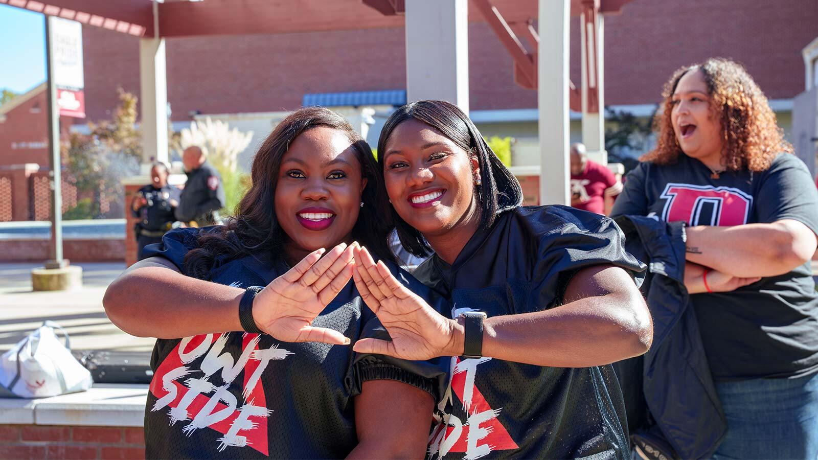 Two smiling women pose together making hand signs in front of a covered outdoor courtyard.