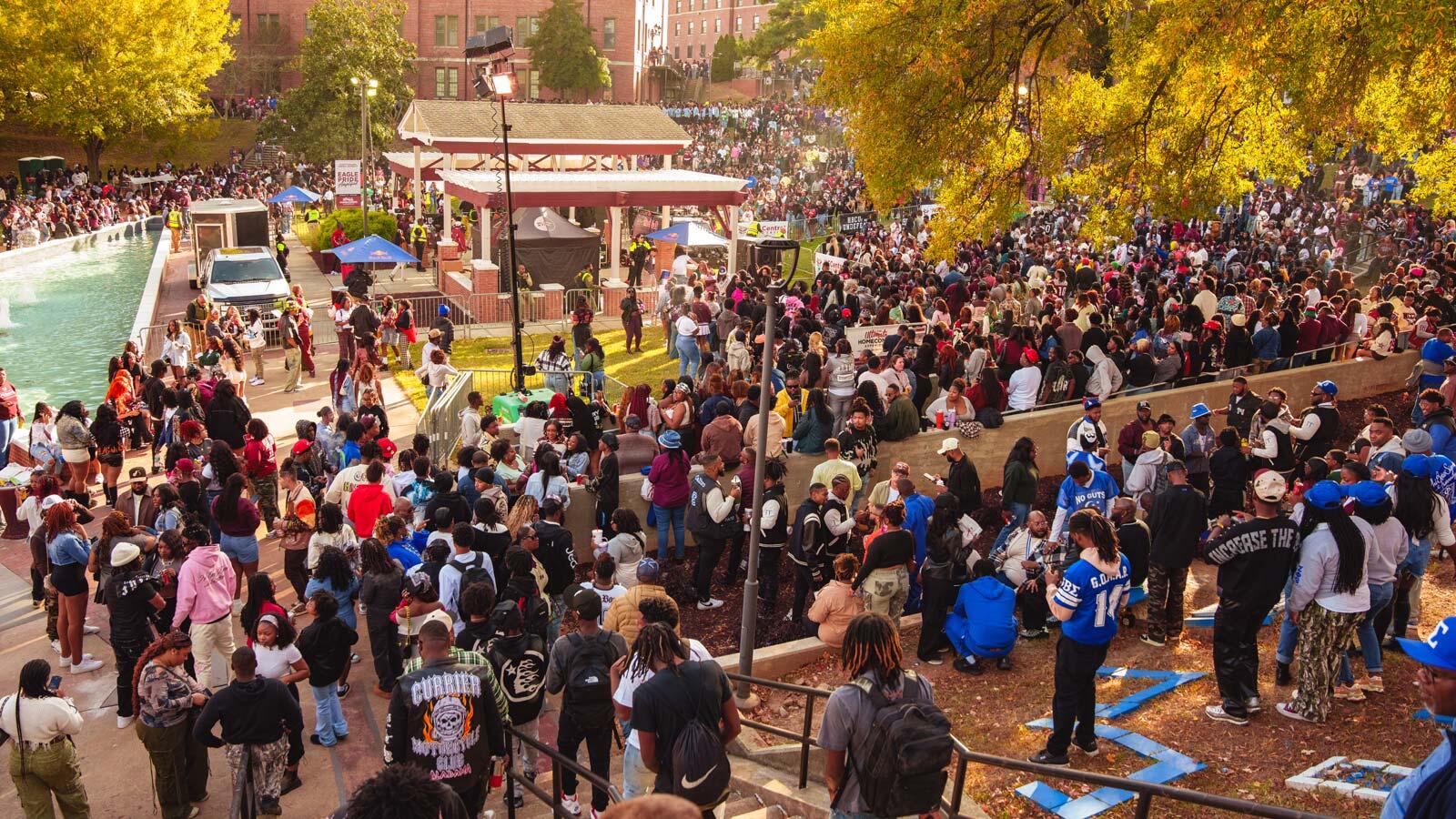 A large crowd gathers around a campus courtyard and reflecting pool surrounded by autumn trees during a university event.