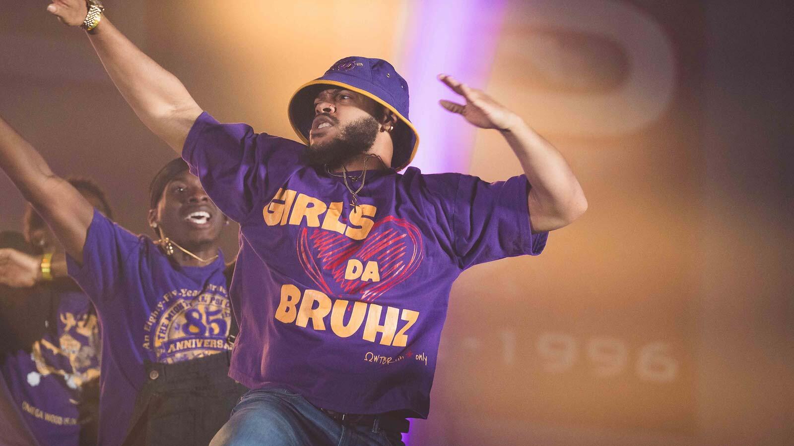 A man wearing a purple bucket hat and a t-shirt reading "GIRLS LOVE DA BRUHZ" performs dynamically on stage alongside other group members