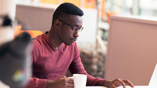 Student with glasses, holding a coffee mug while using a laptop computer
