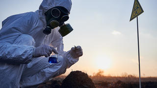 Crouched researcher wearing white protective suit, gas mask and gloves collecting samples from scorched anthill, holding glass flask with blue liquid and tweezers on sunset, near yellow biohazard sign