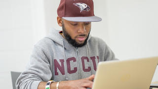 NCCU student on laptop, wearing gray sweater and maroon hat