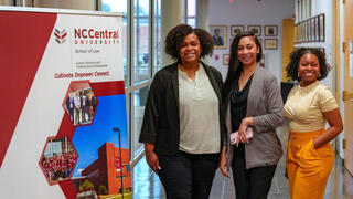 Three women stand smiling next to a banner for NC Central University School of Law.