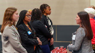 A diverse group of professional women are engaged in conversation, with one woman facing the others and speaking.