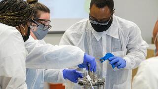  Three individuals, wearing white lab coats, safety goggles, and face masks, are working together on a piece of stainless steel laboratory equipment (likely a bioreactor or similar vessel).
