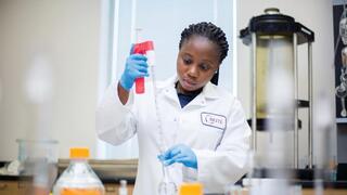  A Black female researcher in a white lab coat (with a "BRITE" logo) and blue gloves is carefully using a pipette in a laboratory setting.