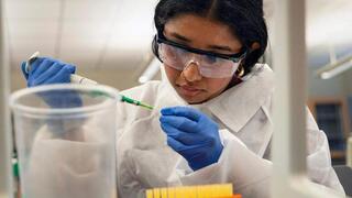  A researcher, wearing a white lab coat, safety goggles, and blue gloves, is precisely pipetting a green liquid into a small tube.