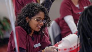  A smiling nursing student with glasses and curly hair is working at a table, wearing a maroon scrub top and clear gloves; her name tag identifies her as a NCCU Nursing Student.