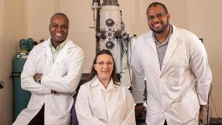 Three smiling researchers, two men and one woman, are posing in white lab coats next to a large scientific instrument (likely a microscope).