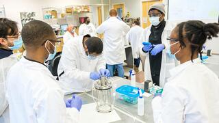 A diverse group of students and an instructor in lab coats, masks, and gloves are collaborating on a piece of bioreactor equipment in a lab.
