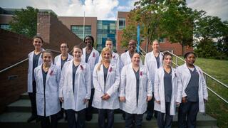 A large group of diverse female students wearing white lab coats and scrubs are posing on the exterior steps of a college building.
