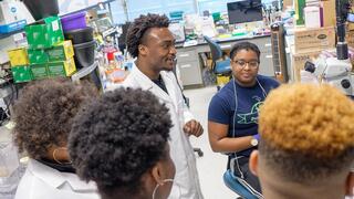  An African American male researcher in a lab coat is leading a discussion with three students in a busy laboratory.