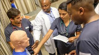 A group of four medical/nursing students and instructors in scrubs and a lab coat are training around a mannequin in a clinical simulation room.