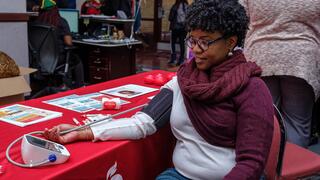  A woman with glasses and a burgundy scarf is having her blood pressure taken at a table with health information.
