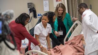  A diverse group of nursing students and instructors are gathered around a patient in a hospital bed (likely a mannequin for training) in a simulated training room.