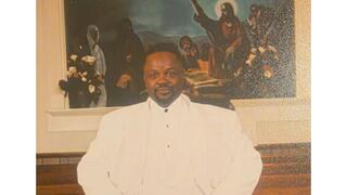 Portrait of Mr. James Winston wearing an all-white suit, standing confidently inside a church sanctuary with wooden pews and religious artwork in the background.