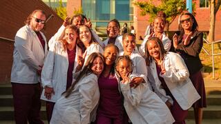 Group of diverse healthcare students in white lab coats smiling and posing together outside a university building.