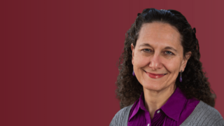 Woman in gray sweater and purple shirt smiling. On maroon background.