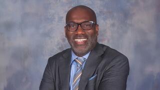 Professional headshot of Joseph Karl Grant smiling, wearing glasses and a suit with tie against a neutral studio background.